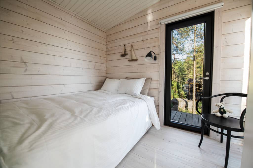 Pieni, valoisa makuuhuone, jossa on vaaleat puuseinät, valkoinen sänky, musta tuoli, jossa on pehmolelu, ja lasiovi, joka johtaa ulos metsänäkymiin.