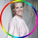 Hymyilevä nainen vaaleanpunaisessa puserossa seisoo kuvioitua seinää vasten, jota kehystää pyöreä sateenkaarenvärinen reunus - tämä muistuttaa Nina Honkasen muotokuvissa nähtyä eloisaa tyyliä.