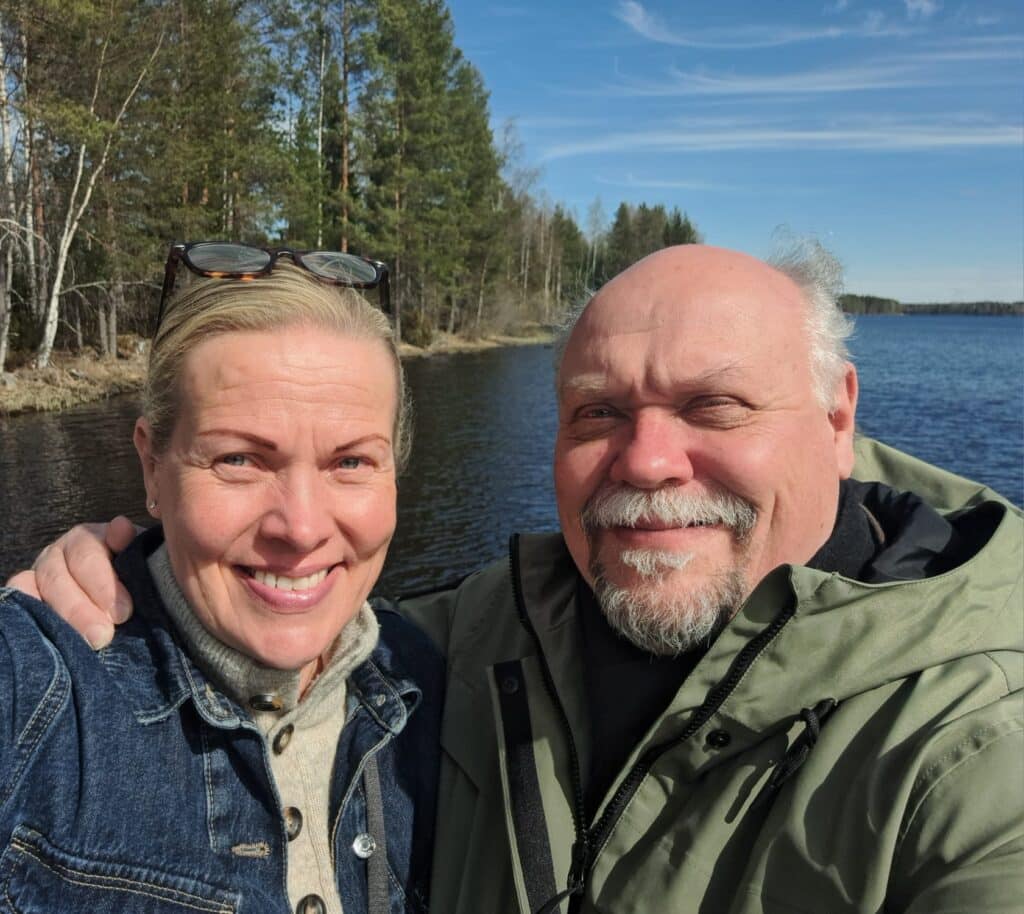 Kaksi aikuista, mahdollisesti Hannu-Pekka Björkman ja Nina Honkanen, hymyilevät ja poseeraavat selfietä ottaessaan ulkona järven rannalla, taustalla puita ja osittain pilvinen taivas, ehkä nauttien mökkiprojektistaan.
