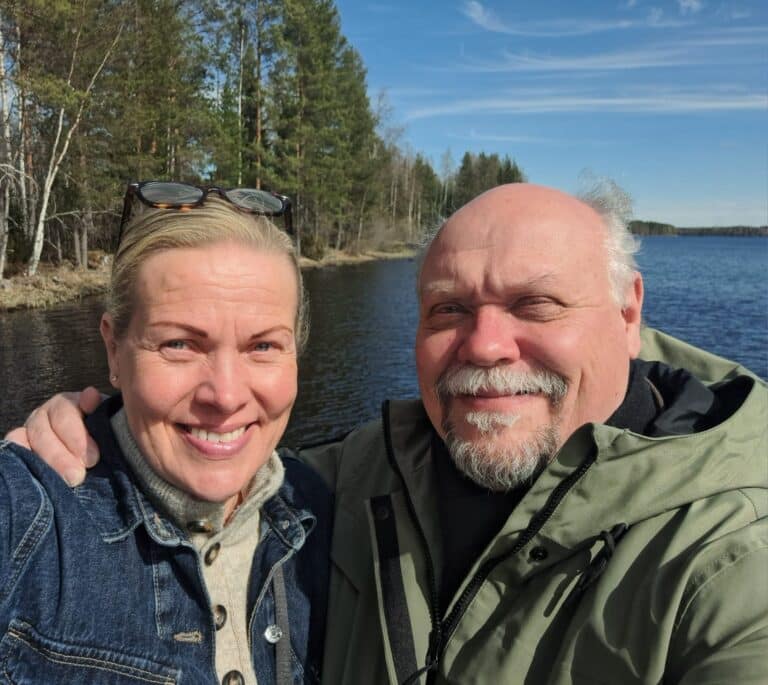 Kaksi aikuista, mahdollisesti Hannu-Pekka Björkman ja Nina Honkanen, hymyilevät ja poseeraavat selfietä ottaessaan ulkona järven rannalla, taustalla puita ja osittain pilvinen taivas, ehkä nauttien mökkiprojektistaan.
