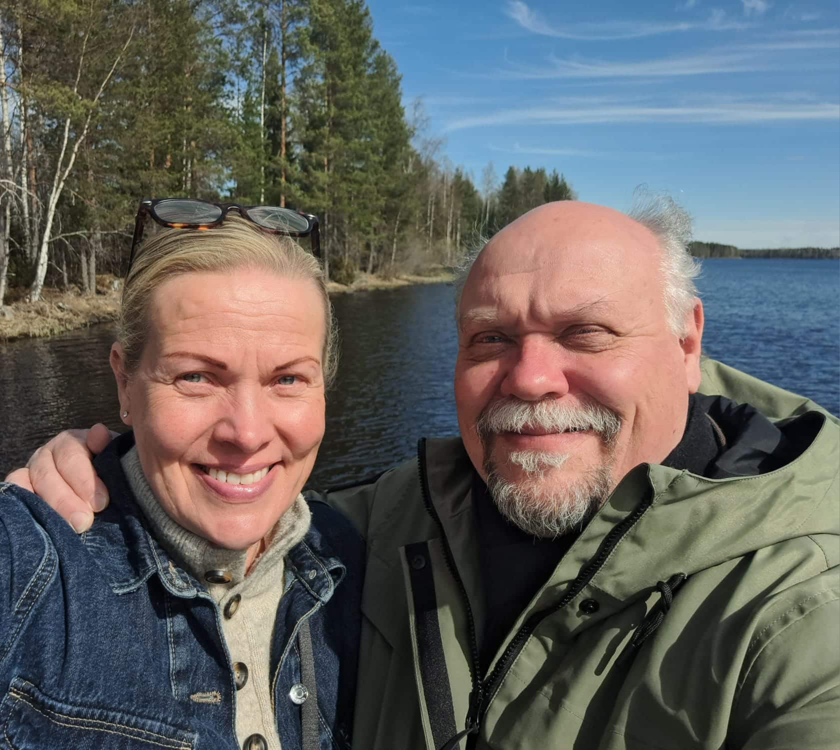 Kaksi aikuista, mahdollisesti Hannu-Pekka Björkman ja Nina Honkanen, hymyilevät ja poseeraavat selfietä ottaessaan ulkona järven rannalla, taustalla puita ja osittain pilvinen taivas, ehkä nauttien mökkiprojektistaan.
