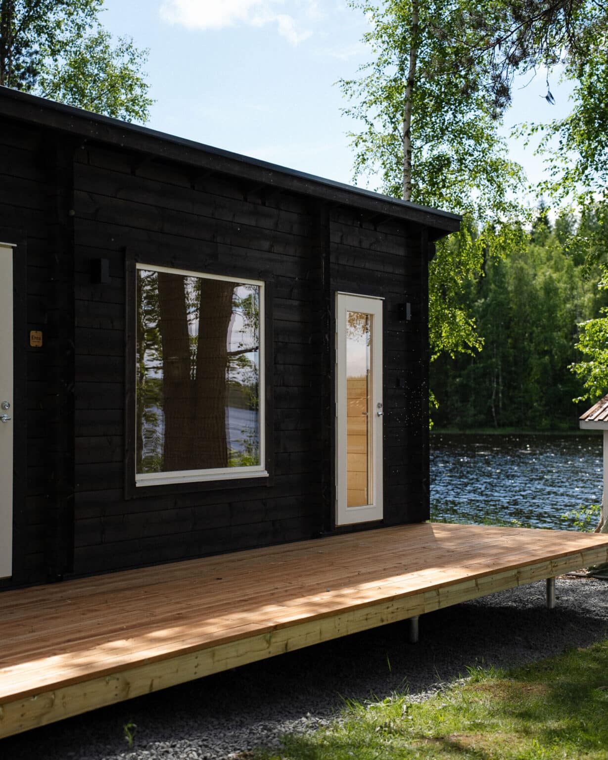 Moderni musta mökki, jossa on suuret ikkunat ja puuterassi, avautuu järvelle, ja sitä ympäröivät puut ja vehreys aurinkoisena päivänä.