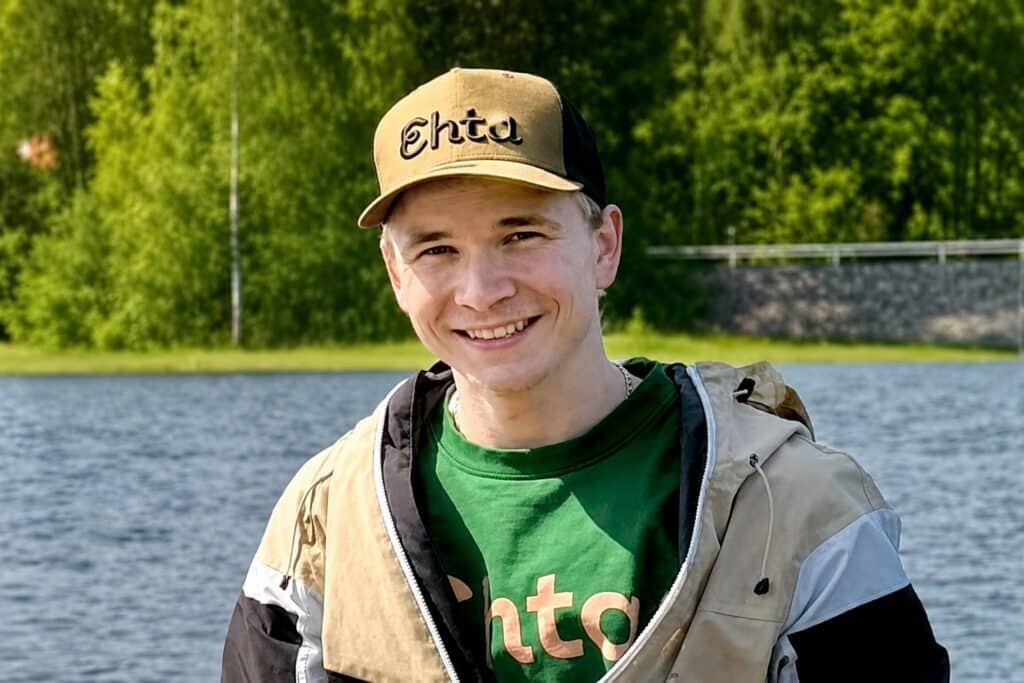 Hymyilevä henkilö, jolla on päällään ruskea ja musta lippalakki sekä vihreä paita, seisoo ulkona järven rannalla; taustalla näkyy vihreitä puita ja aita. Ota yhteyttä, jos haluat lisätietoja tästä viehättävästä paikasta.