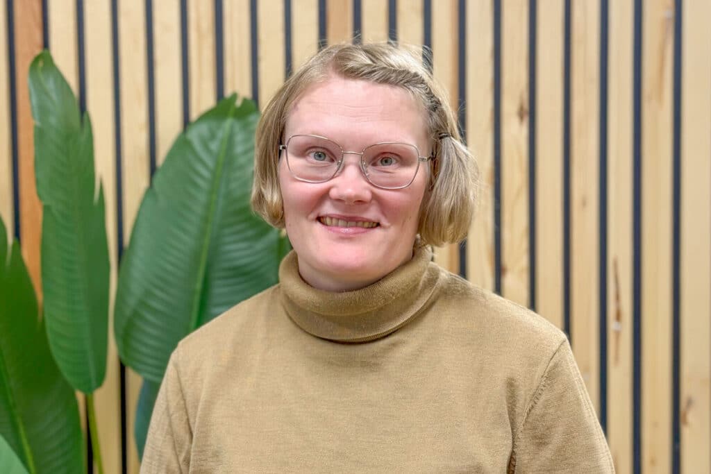 Lyhyttukkainen, vaaleahiuksinen ja silmälasit päässään oleva henkilö, jolla on yllään vaaleanruskea turtleneck-villapaita, seisoo sisätiloissa kasvin ja puisten säleiden muodostaman seinän edessä. Lisätietoja tai kyselyitä varten ota yhteyttä.