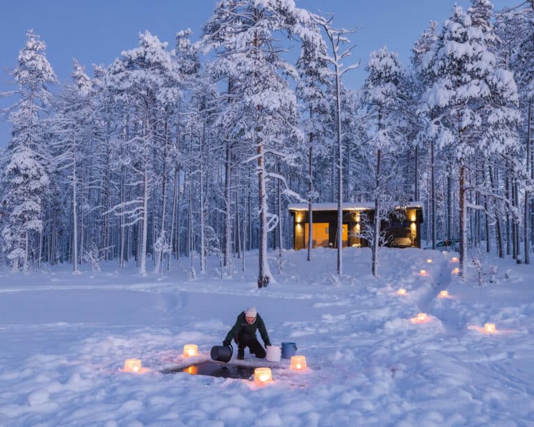 Hutun saunamökki lumisessa männikössä illalla, avannolle johtaa lyhtyjen rivi