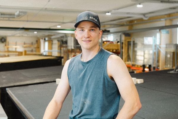 Tumma lippalakki ja hihaton paita päällään oleva saunanrakentaja seisoo rakennustyömaalla sisätiloissa, työkalut vyöllään ja rakennusmateriaalit taustalla.