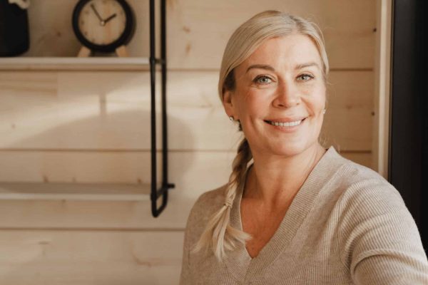 Nainen, jolla on vaaleat hiukset letissä, hymyilee kameralle huoneessa, jossa on vaalea puupanelointi, joka kuvastaa saunamökin suunnittelussa usein pidettyä kodikasta tyyliä, ja taustalla on kello hyllyllä.