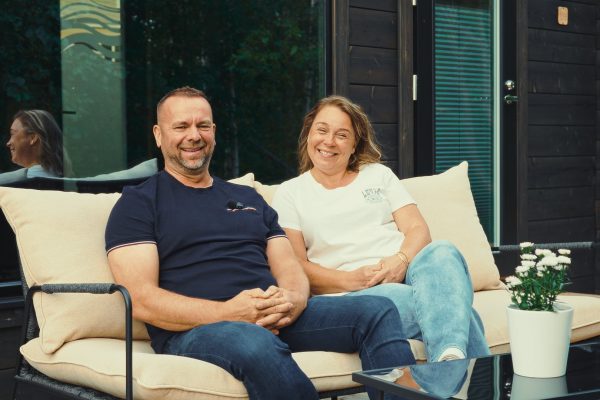 Mies ja nainen istuvat hymyillen ulkosohvalla pienen pöydän vieressä, jossa on ruukkukasvi, ja rentoutuvat valmis saunamökin edessä, jossa on tummat puuseinät ja lasiovet.